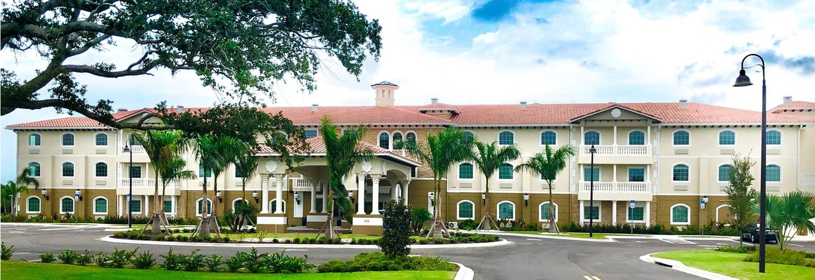 Front view of Chateau Madeleine with a Mediterranean-style senior living community with three stories, light beige and tan exterior, red-tiled roof, white columns at the entrance, and multiple balconies.
